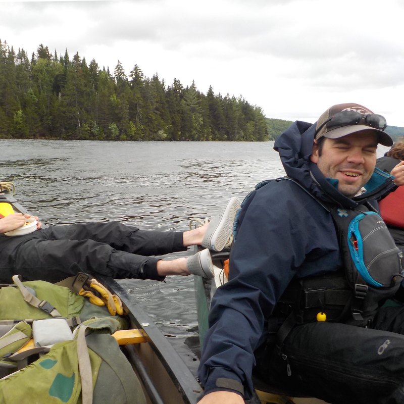 The image shows two people in a canoe on a lake. One person is wearing a blue jacket and a life vest, and the other is lying down with their feet up. The background features a forest and a cloudy sky. The overall scene suggests a relaxing day on the water.
